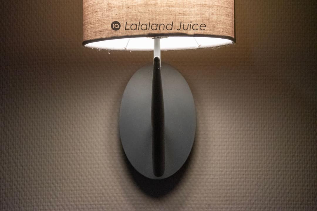 A single warm wall lamp glowing against a textured wall in a dimmed room — the Lalaland Juice mark set within.