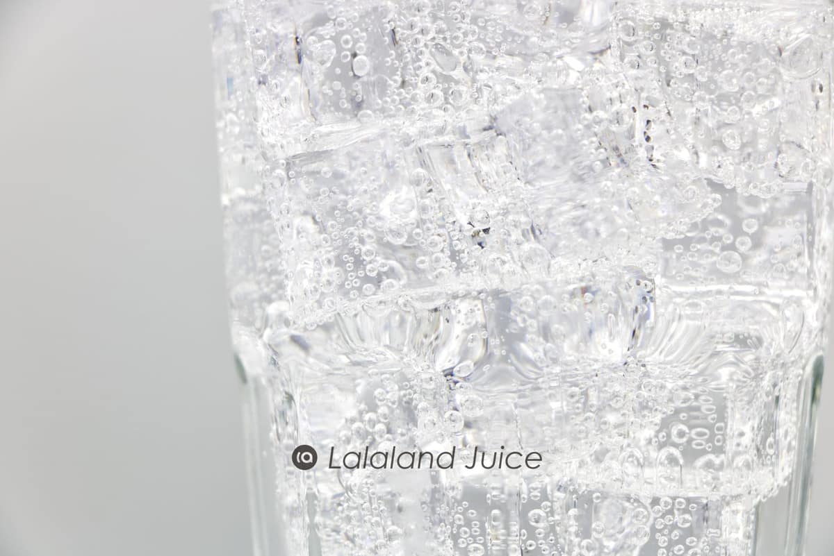 A tall glass packed with ice and sparkling water in close-up, beaded and cold — the Lalaland Juice mark set quietly within.