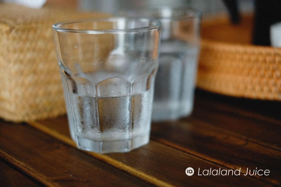 A cold glass of water beaded with condensation on a rustic wooden table — water first, before the kopi.