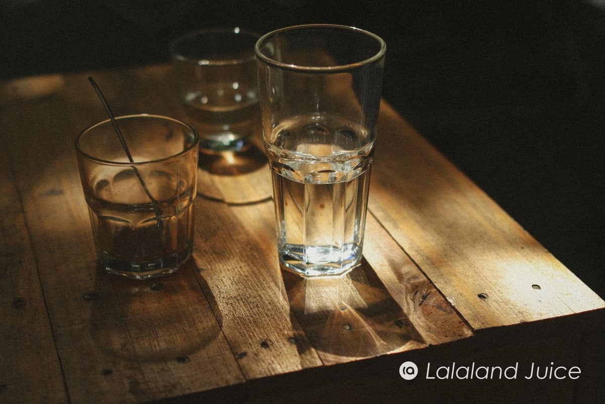 A tall glass of water catching a shaft of warm morning light on a wooden table — the first small kindness of a gentle morning — with the Lalaland Juice mark set within.