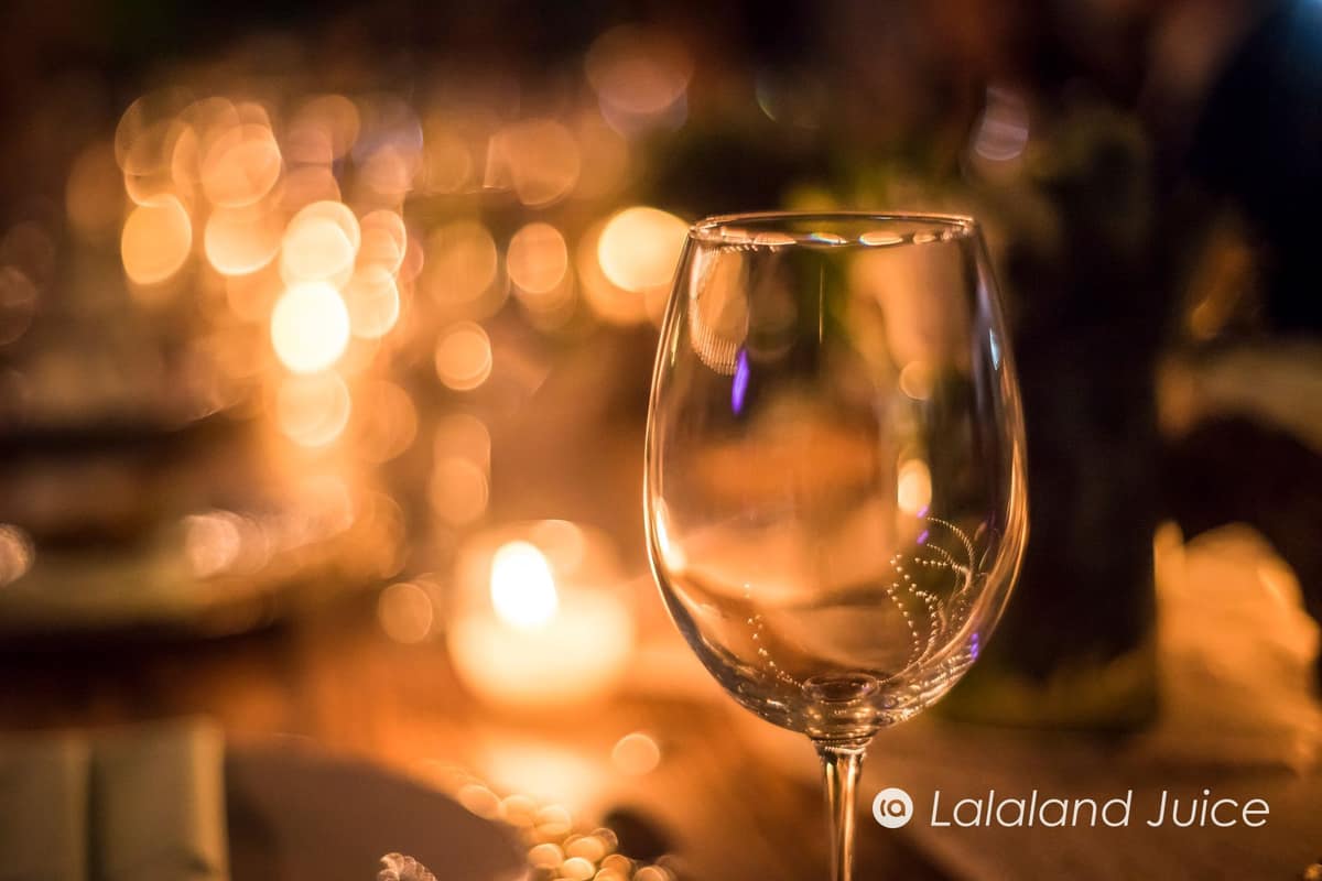 An empty stemmed glass at a candlelit table, the warm lights of the evening softened behind it — the Lalaland Juice mark set quietly within.